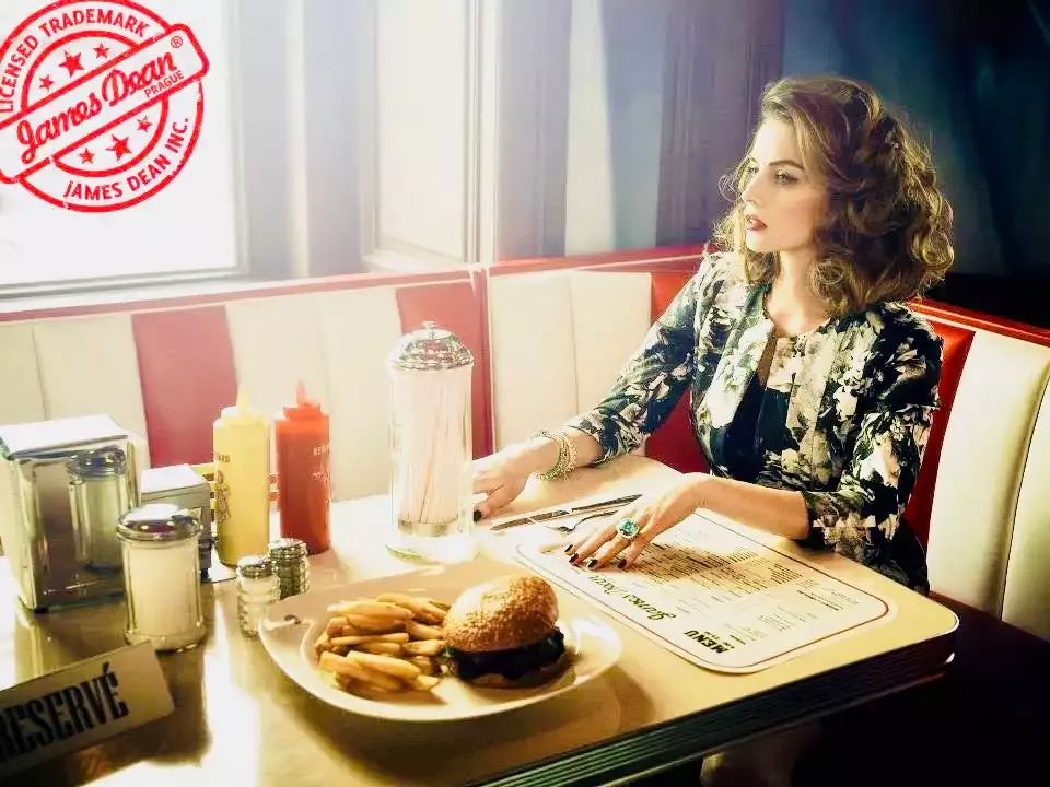 Modelka u okna v boxu dineru, na stole burger s hranolky, kečup a hořčice