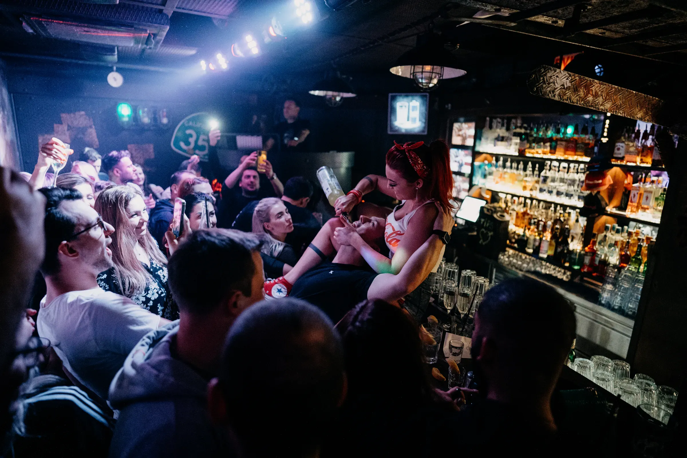Tanečnice v tílku Coca-Cola na baru Music Clubu a hosté, kteří ji natáčejí telefony
