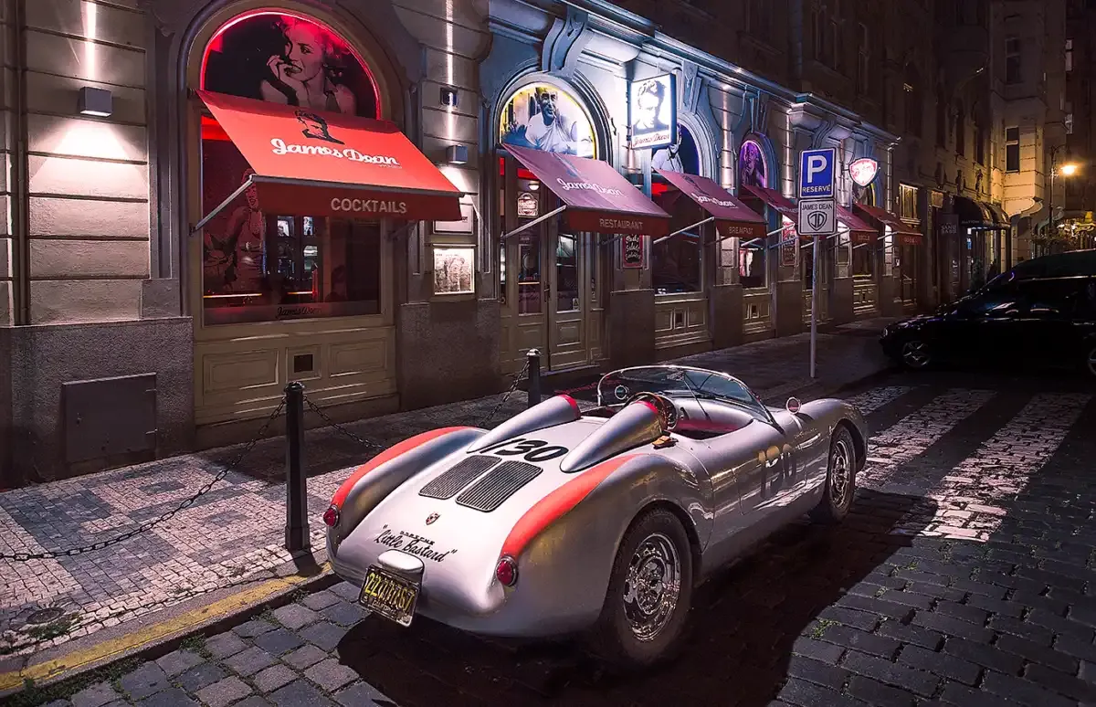 Noční průčelí James Dean Prague s červenou markýzou Cocktails a stříbrný sportovní roadster z 50. let na dlažbě